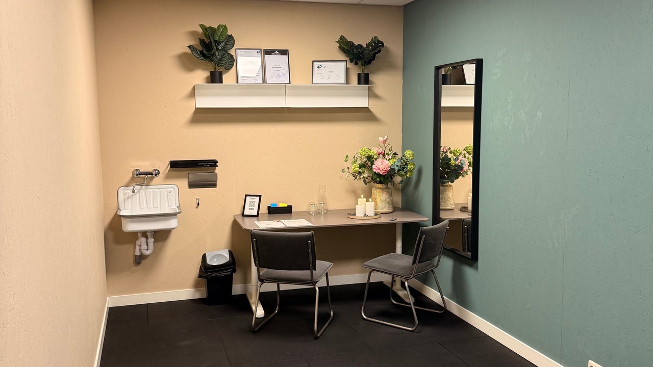 De behandelkamer: wastafel, bureau met bloemen, planken aan de beige muur en de zwarte spiegel aan de groene muur